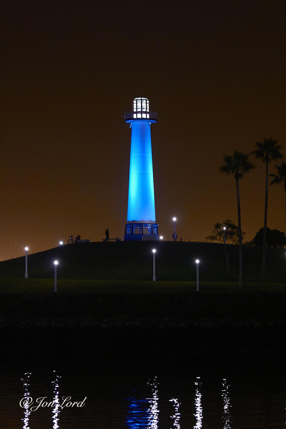 This is a night time colour photo in portrait format of a tall, illuminated lighthouse located on a small hill, harbour water in the foreground and a dark caramel coloured sky in the background. Long Beach, California (2012).

The dark waters of the harbour rise from the base of the photo, upwards to a little less than the one quarter up level, where the boundary between the water an distant shore is ill defined. The camera is located on the near shore, a few metres above the water and angled at the distant shore and lighthouse. The jet black water is rippled with the six long reflections from the small street lights on the shore. One further and shorter reflection is blue from the illuminated lighthouse. Above the shore and just less than the halfway up level is a path that slopes upwards gently from the right to the left. Along this path are four of the cylindrical poles with a light on top. Behind the path is the small hill with the lighthouse on top, in the centre of the photo and 