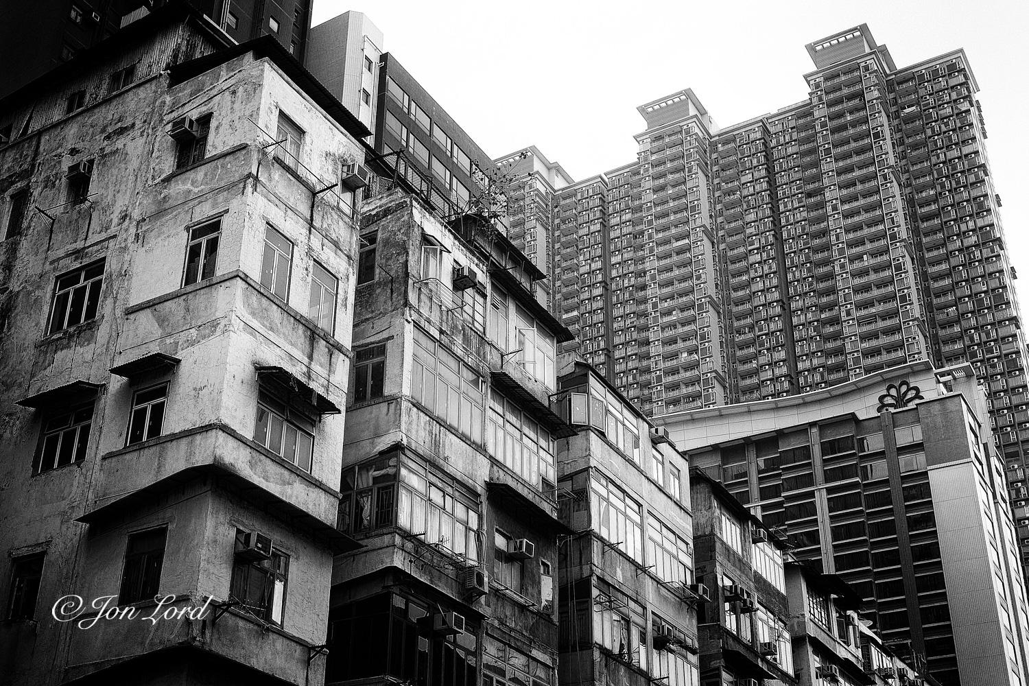 This is a black and white cityscape photo in landscape format of an old and weathered low-rise apartment building in the foreground and a very high-rise modern apartment building in the background. To Kwa Wan, Hong Kong (2017). 

In the foreground, stretching from the left edge of the photo diagonally downwards to the lower right corner, is the front facade of our mid twentieth century apartment block. In reality it's probably mixed use with businesses intertwined with residential spaces. The camera is located on the opposite side of the street, angled upwards and viewing along the front of the facade which appears to be made up of six or more closely spaced buildings with the ground floors hidden beneath the photo. Each block is heavily weathered and once may have been painted white. As is common with this era of building, the types of window, location of external A/C units and drying washing - is totally random. The buildings are low-rise being limited to a maximum of ten floors due 