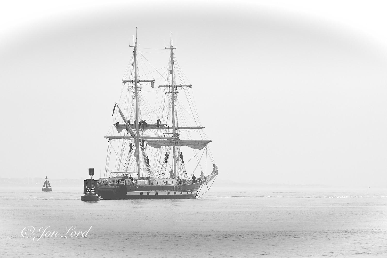 This is a black and white photo in landscape format of a Brig (square rigged tall ship) underway through the deep water channel with the crew on the yards setting sail. Southampton Water, UK (2019).

The near calm waters of the Solent rise upwards from the base of the photo to the quarter up level. The horizon is ill defined since there is an early morning sea fog reducing the visibility to less than one km. In the lower left corner of the image are two large navigation buoys, the closer dark buoy sits on a large cylindrical base with four tall vertical, rectangular panels on the sides and a square panel at the top. A little further away is a further buoy, lighter in tone than the first, with four panels that slope inwards creating a triangle: topped with a further triangle. Very slightly to the left of centre in the photo is our square rigged tall ship that has just passed between the two buoys from left to right. The camera is located two metres above the water with the vessel being 