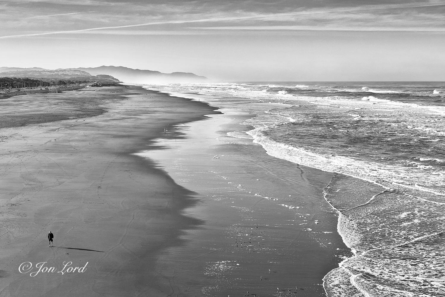 This is a black and white photo in landscape format of an elevated view along a nearly deserted and wide beach that stretches for miles with the sea on the right side and a single man walking in the lower left corner. Ocean Beach, California (2017).

The camera is located thirty metres above the beach and is aligned with the beach on the left side of centre and the sea on the right. The beach and ocean rise upwards from the base of the photo to a little over the three-quarters up level. The beach is wide, at least one hundred metres from the shore to the water, perhaps more. The beach has ragged streaks extending along parallel to the water, indicating the high water mark and the point where the surf from the breaking waves is reaching: the latter has a shiny surface from the recent passage of water. From the centre to the right edge of the image is the Pacific Ocean. The beach must continue out into the sea with a gentle and shallow gradient since there is one hundred metres of surf b