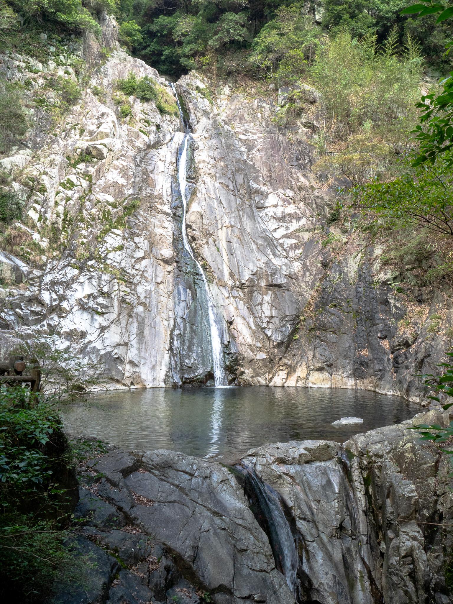 A tall but narrow waterfall spilling down a steep cliff of light brown and gray rock, with a scattering of green plants. Above the waterfall are some trees. The waterfall spills into a round pool of water. At the front of the pool a second waterfall begins in the foreground. To the left of the pool are some leafy plants and a wooden structure right at the edge of the frame. The main waterfall is well lit by the sun, but the foreground is in shade.