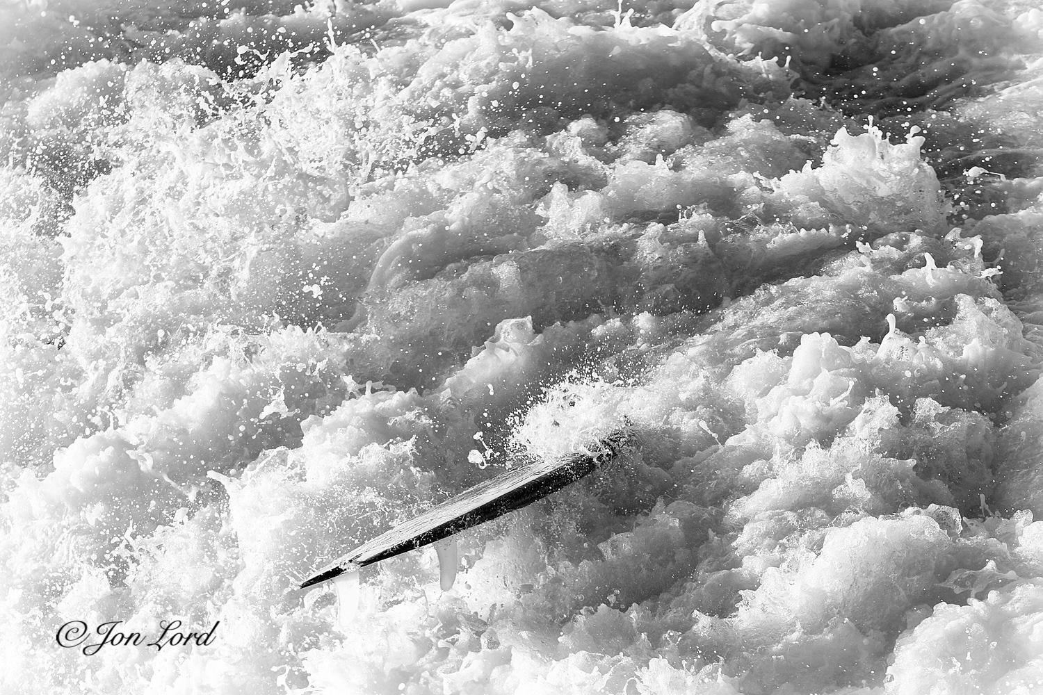 This is a black and white water sports photo in landscape format of the rear of a surfboard protruding out of a brilliant white and swirling mass of foaming surf. Hermosa Beach (2014).
The entire image is overflowing with the maelstrom of boiling surf from a breaking Pacific wave. The foaming water is brightly lit by the still low morning sun. Below the centre of the photo is the surfboard. The camera is located a few metres above and a few metres away from the board. Only the back and part of