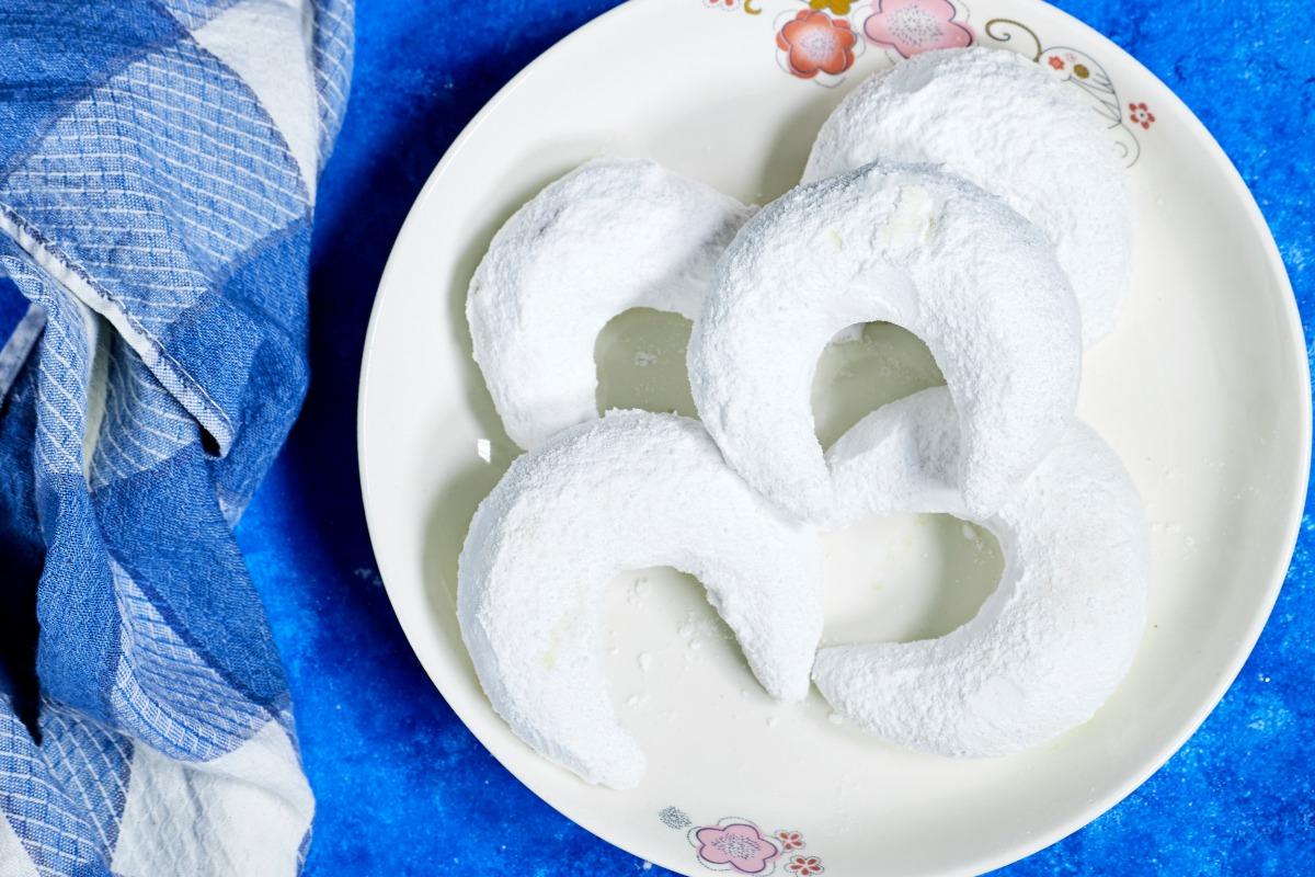 Crescent-shaped gazelle horn almond cookies heavily dusted with powdered sugar on a white floral-rimmed plate against a bright blue background.
