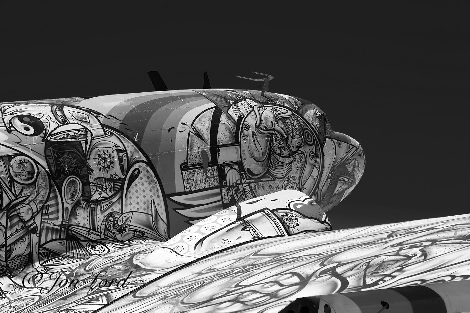 This is a black and white photo in landscape format of an old Douglas DC3 aircraft painted in a psychedelic style. Tucson, Arizona (2013).
The aircraft stretches from the left margin across the photo towards the right edge. The fuselage is angled upwards with only the mid and forward section in view. The wing stretches outwards from the middle of the fuselage and ends out of view beyond the right of the image. The camera is located close behind the wing, a metre and a half above the ground and