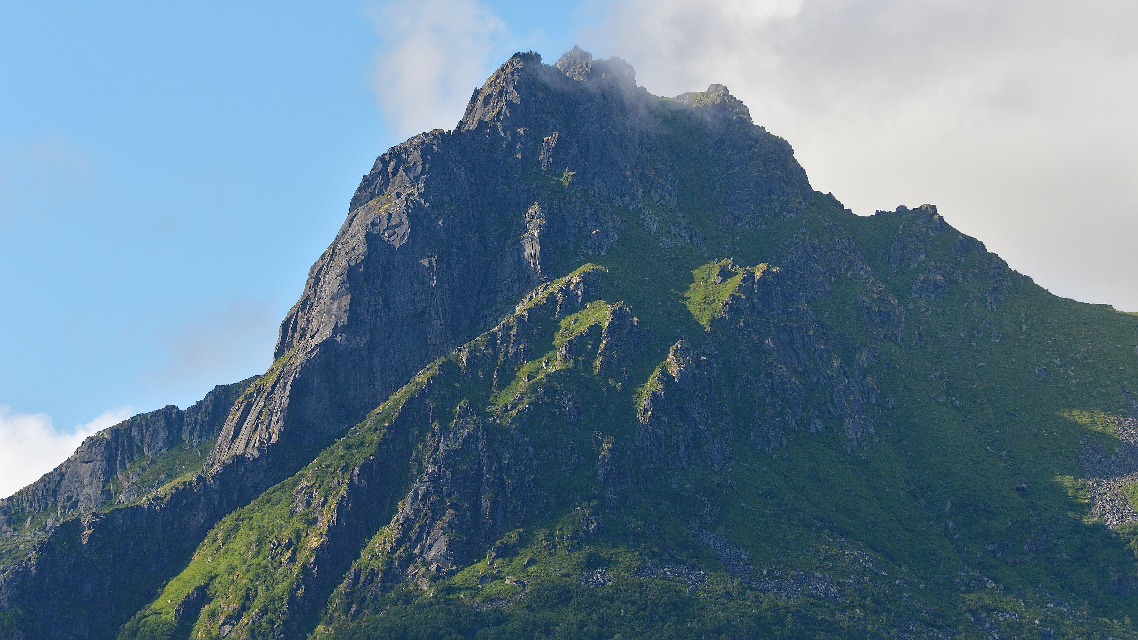 A photo of a green and rocky mountain peak. The sky is blue with white clouds going over the peak.