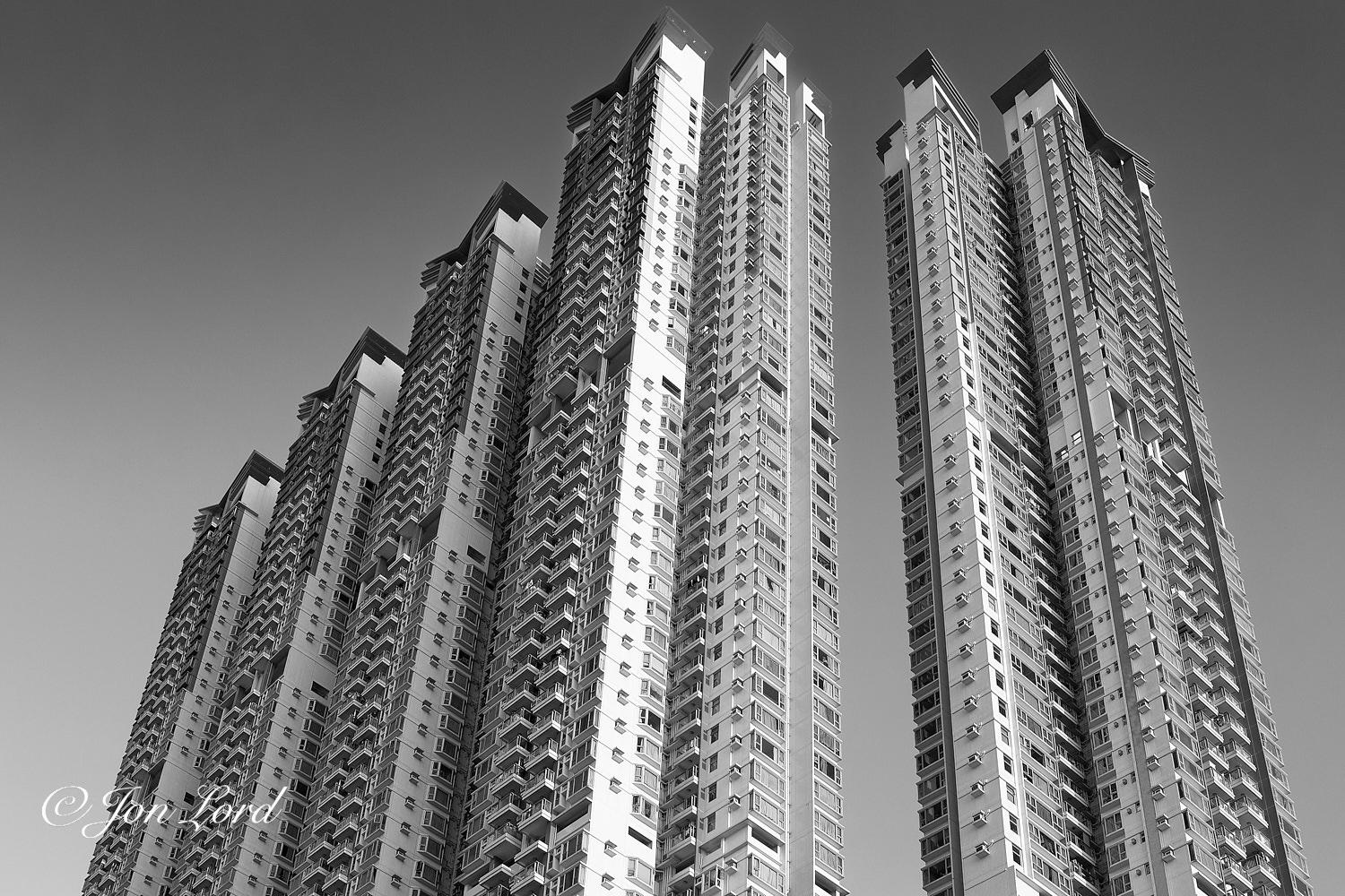 This is a black and white photo in landscape format looking up at five 57 story apartment blocks. To Kwa Wan, Hong Kong (2016).
The camera is set back perhaps fifty metres from the buildings and angled upwards steeply so that the lower floors are hidden below the lower edge of the photo and no other buildings or objects are in view. While there are five blocks: each separate block appears as two blocks that are joined together at a corner. The blocks have a rectangular footprint and are tall an