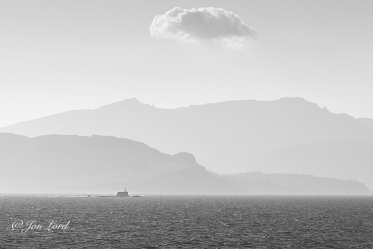 This is a black and white photo in landscape format of a modern submarine running on the surface with rising terrain in the background. Firth of Clyde (2017).

The dark waters of the sea rise from the base of the photo upwards to a little less than the quarter up level. On a poorly defined horizon and below and to the left of centre is our submarine, underway from left to right and just above the surface. The camera is located a few metres above the water with the submarine being around one km away. The all black vessel is viewed in profile with the starboard (right) side in view. Water can be seen washing over the casing (top of the hull) with a short sensor above the deck aft of the bow. Further aft is the large fin (sometimes called the sail or conning tower) that's located a little forward of midships with two or three raised signal masts, radars or periscopes. Further aft is more dark hull that is faired downwards towards the stern with a short stretch of water before the upper ru