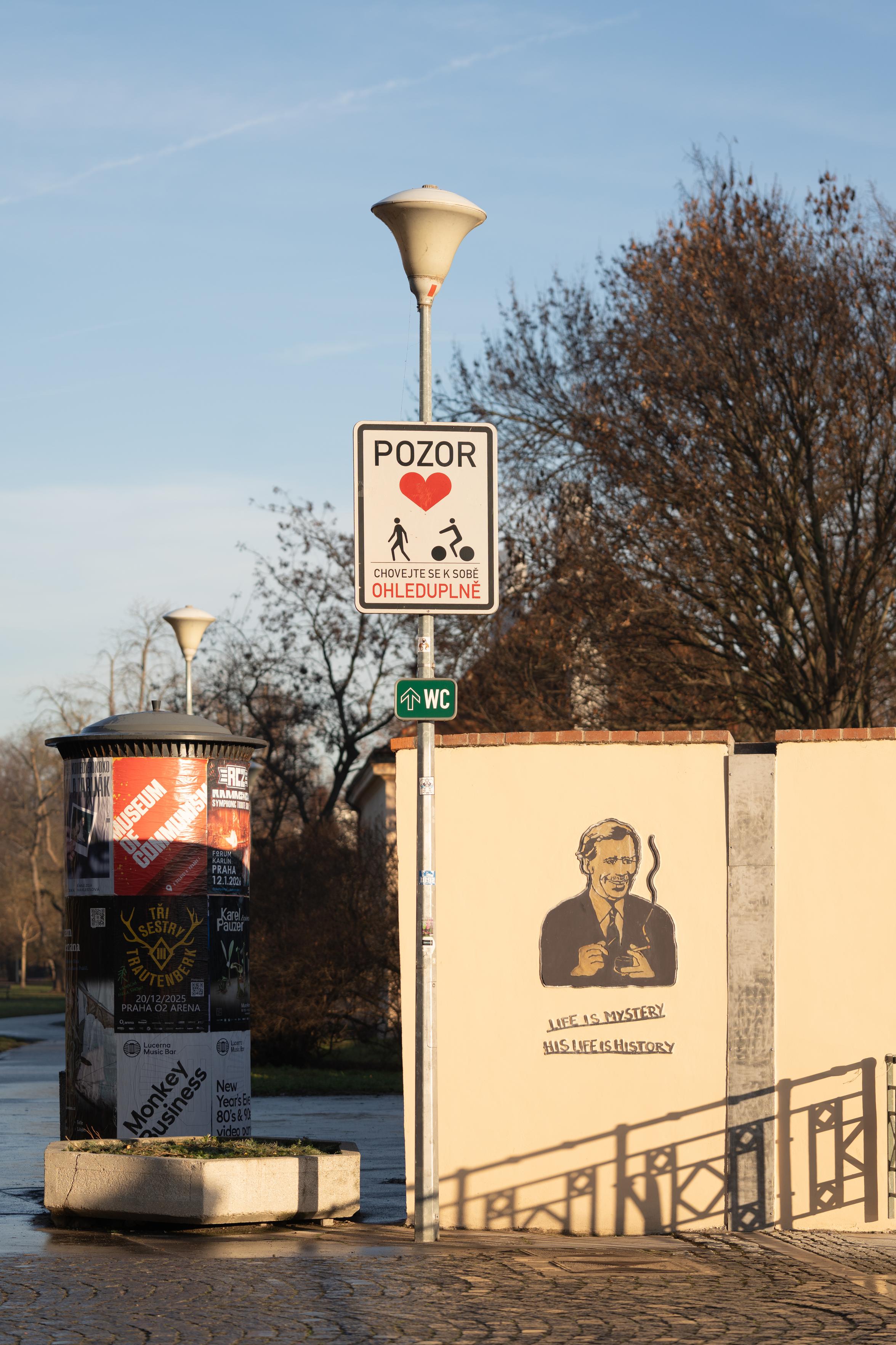 a photo of Prague in morning light that casts a golden glow. a park near the Vltava. an advertising column on the left with various posters. a lantern with a big sign in the midde: POZOR ('caution'), above a big red heart and pictographs of a pedestrian and a cyclist. the sign  asks to be considerate to one another. a small toilet sign underneath. prominently on a wall on the right an artwork of Vaclav Havel and the words "Life is mystery, his life is history"