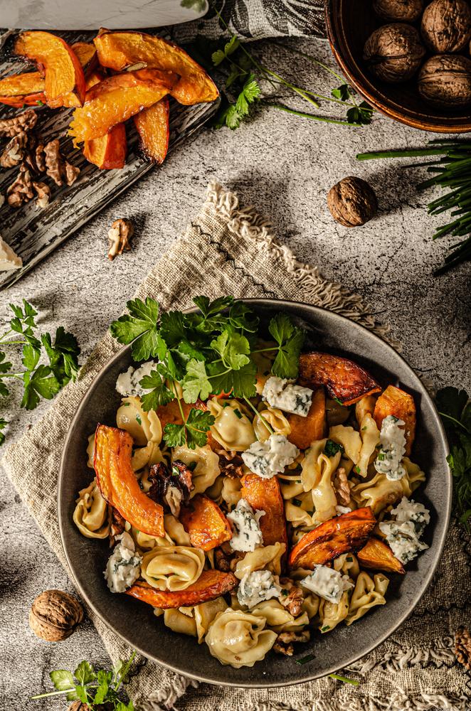 A rustic bowl of tortellini tossed with roasted pumpkin wedges, walnut pieces and crumbled blue cheese, garnished with fresh parsley on a linen napkin and textured stone surface.