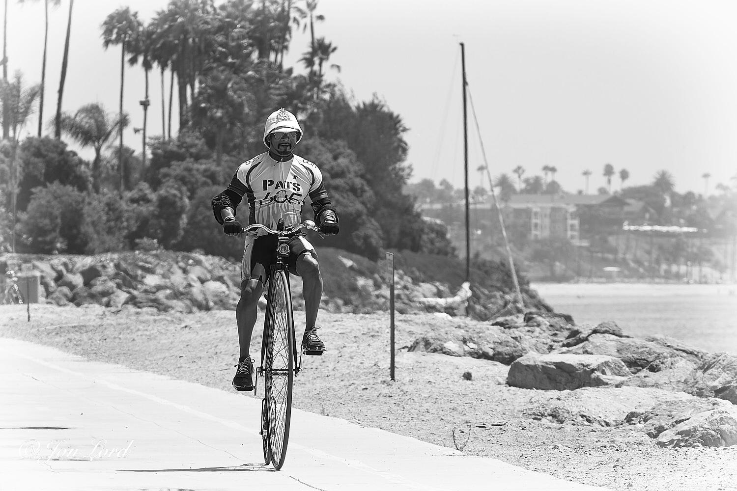 This is a black and white street photo in landscape format of a man riding a Penny Farthing along a waterfront bike path. Long Beach California (2014).

From the lower right corner is the white concrete bike path that extends diagonally up and across the image to the left edge. To the left of centre is our cyclist riding his penny farthing, sometimes called a high wheel. The camera is a metre and a half above the ground, aligned towards the cyclist at a distance of three metres and with the cyclist filling three-quarters of the height of the image. The cyclist is in motion from left to right, diagonally across the photo and towards the camera. The intrepid rider is wearing a white pith helmet, dark aviator style sun glasses, has a short and neat dark Van Dyke beard and is clothed in a cyclist's jacket, shorts and trainers. He also wearing a pair of stout black gloves and elbow pads. To the right of the bike path is a metre or two of sand before a rocky seawall is reached with the ocean