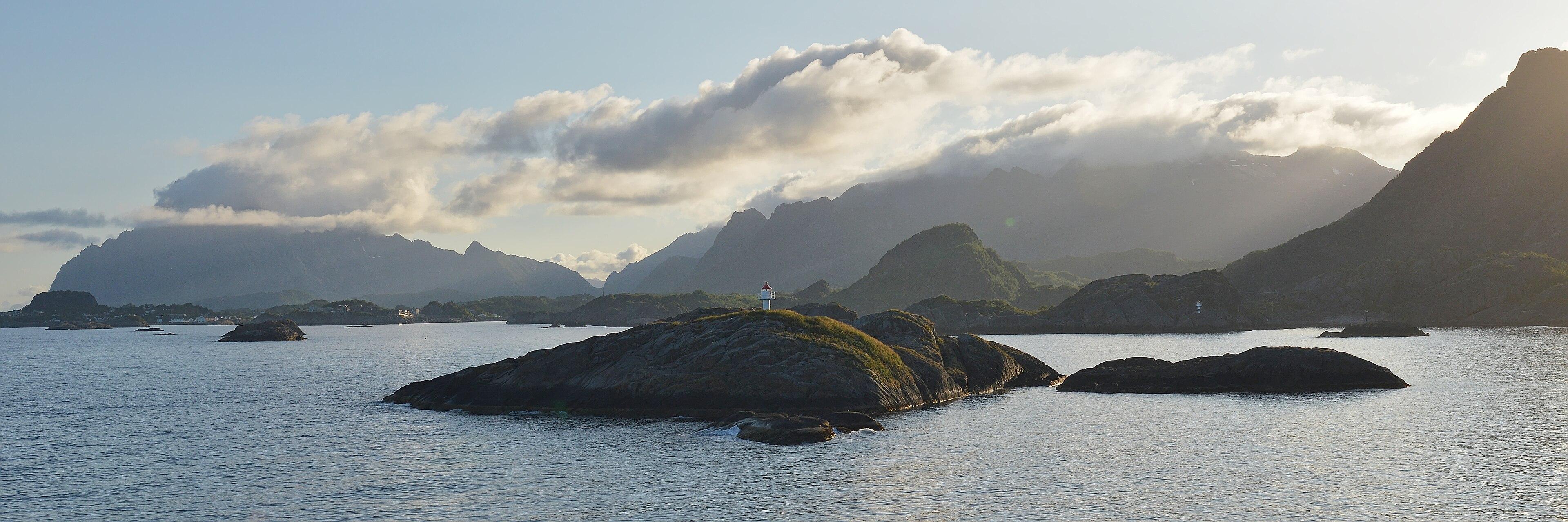 A panoramic photo of small islands in front of mountains. The sky is clear with some cloud going over the mountain peaks.