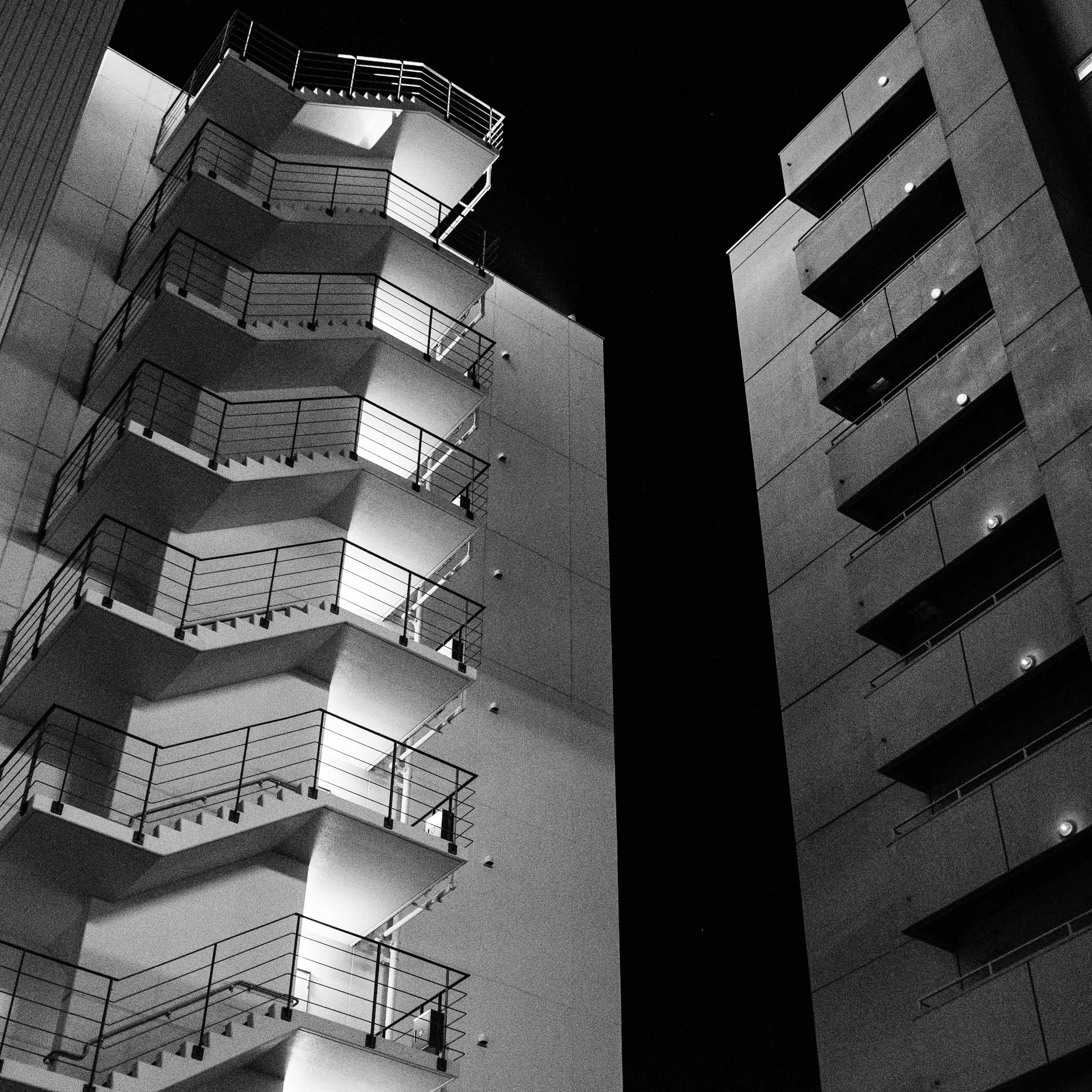 A low-angle night view of two buildings. The left building features a zig-zagging exterior staircase that is brightly illuminated, casting sharp shadows and highlighting horizontal metal railings. The right building has stacked balconies, each lit by a single round wall lamp. The sky between them is pitch black.