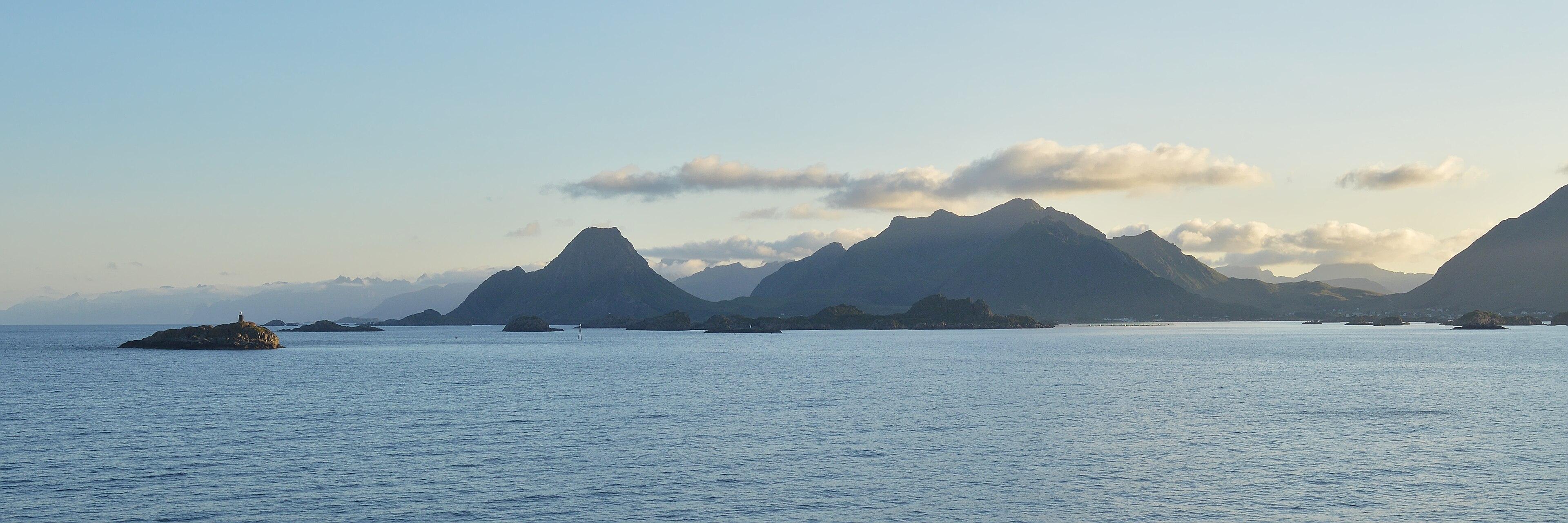 A panoramic photo of small islands in front of mountains. The sky is clear with some low clouds in it.