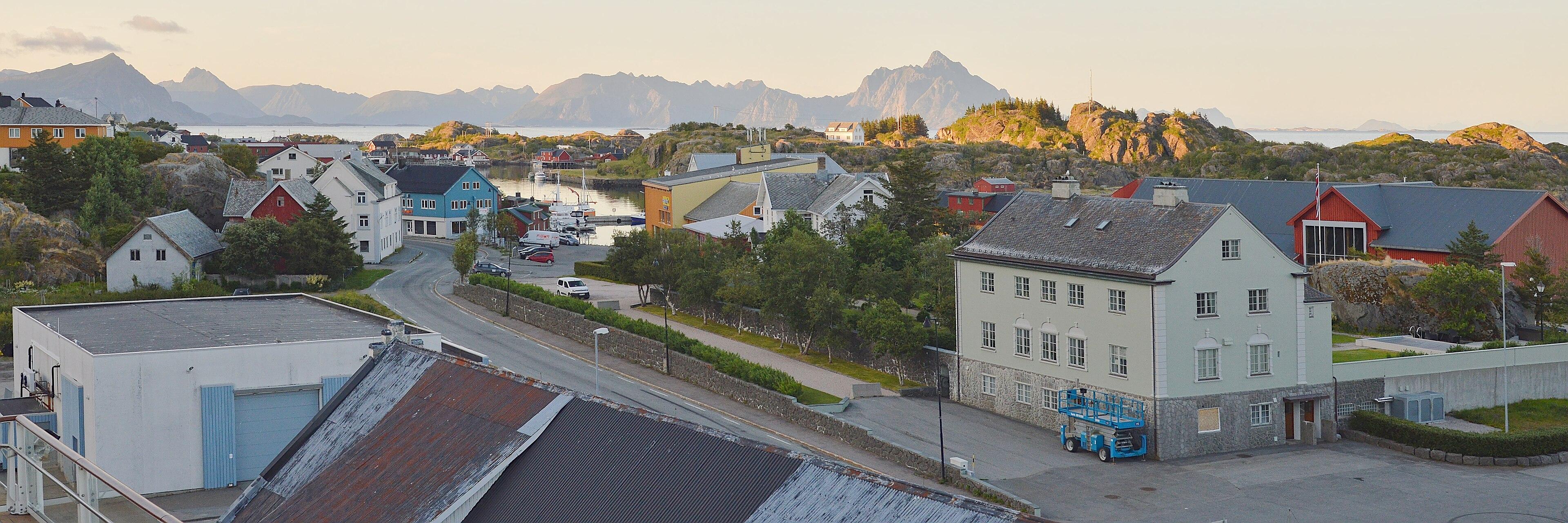 A panoramic photo of the town of Stamsund in Norway. The light is warm and the town is in the shadow of a mountain. There are mountains across the water in the distance. The sky is hazy and clear.