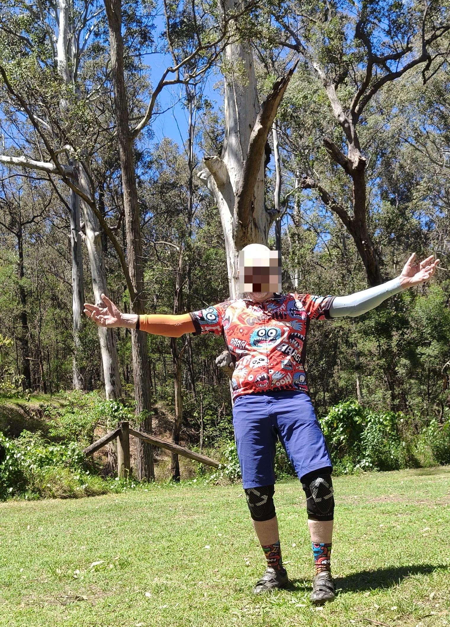 A mildly overweight old bald man stands on grass with his arms outstretched in parkland. Behind is natural bush featuring massive gum trees. It's a gloriously sunny blue sky day.
He is dressed... colourfully. Some would say a mishmash of colours, others a riot, maybe a kaleidoscope or a cacophony, even a rainbow. A vomit?
Our model wears a multicoloured cycling shirt from AliExpress (AE), featuring colourful, scared monsters, broken bones, and cries of pain, reflecting the model's riding histo