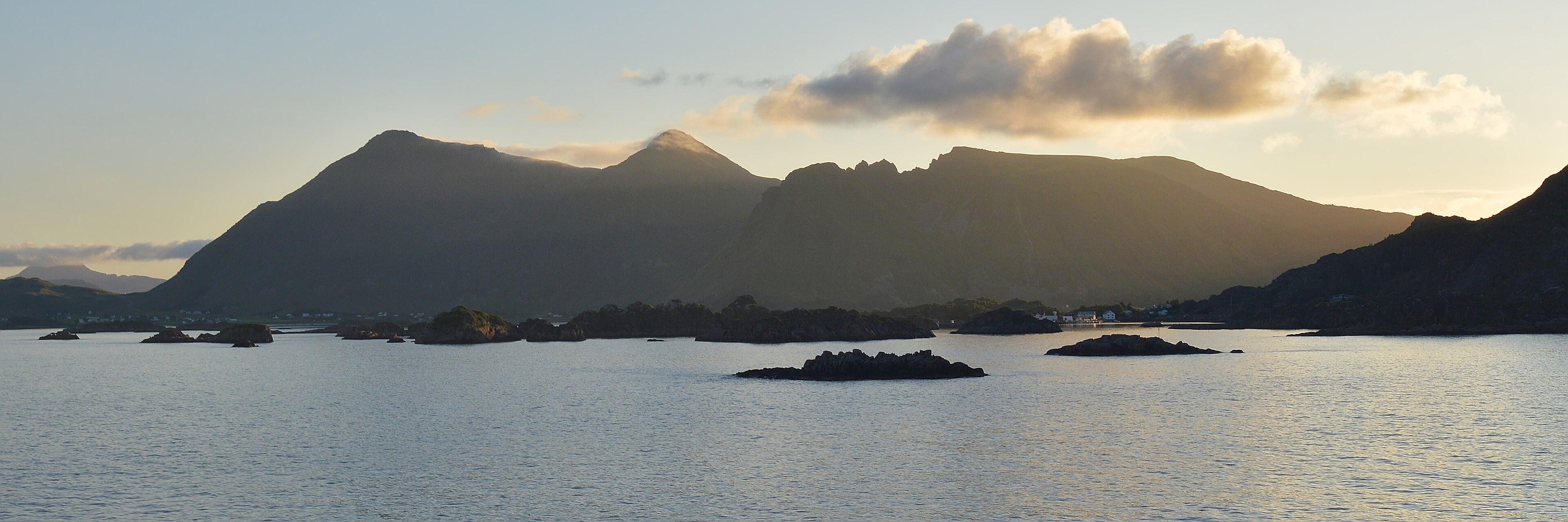 A panoramic photo of mountains with small islands in front of them and the sun behind them. The sky is mainly clear with a few low clouds in it.