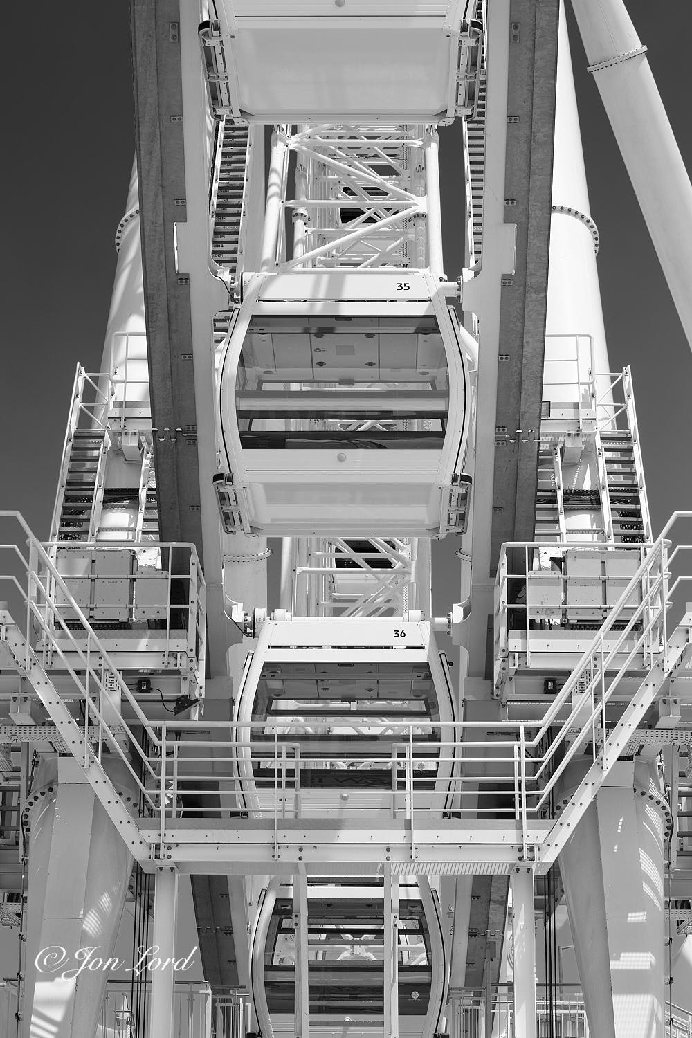 This is a close-up abstract black and white photo in portrait format of a modern city centre ferris wheel. Central District, Hong Kong (2015).
The entire photo is filled with this bright white engineering structure. The camera is located a metre and a half above the ground, elevated upwards slightly, a few metres away from the closest point of the wheel and aligned towards the front with neither side in view. In the foreground and at the base of the photo is a framework of white metal stairs, s