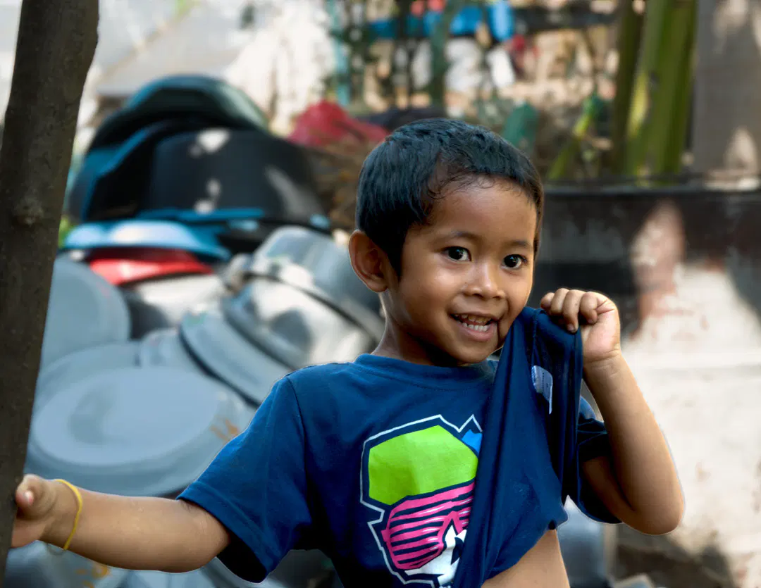 Young boy with short dark hair smiles brightly at the camera. He wears a navy blue t-shirt, pulled up to his cheek on one side; it has a colorful geometric design in green, pink, and white. Blurred pots and pans, and outdoor scenery visible in background.