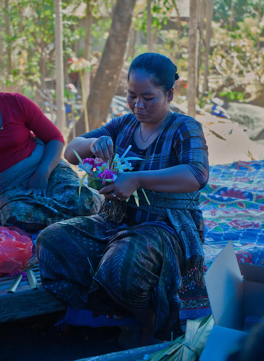 Woman in traditional blue patterned clothing sits cross-legged on colorful woven mats, carefully arranging purple and white flowers with green leaves. She works outdoors under trees with another person in red visible to her left.