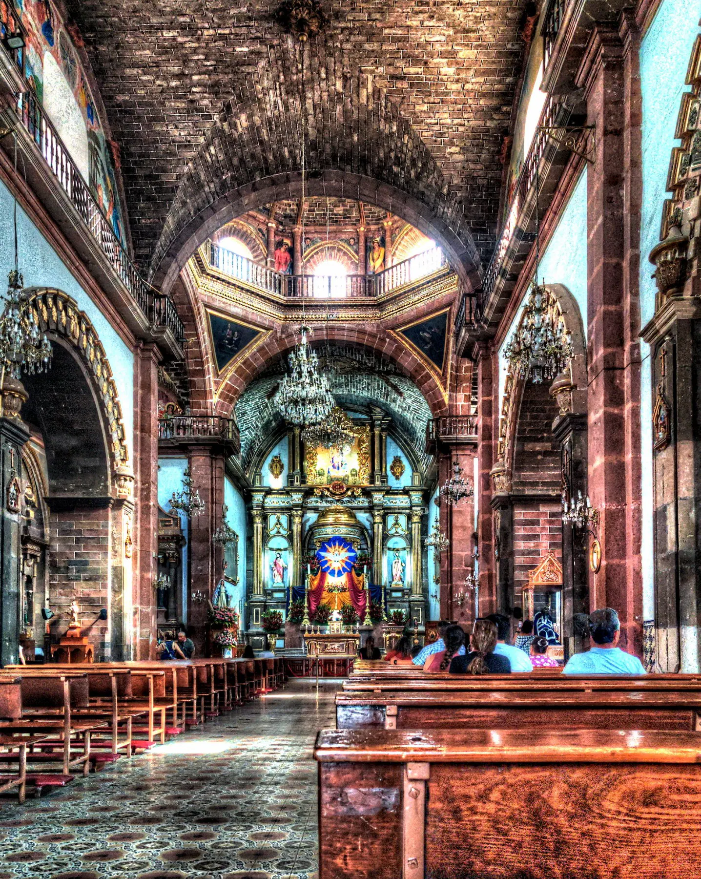 HDR-processed interior of an ornate Catholic church, viewed from the nave looking toward the altar. Wooden pews line the aisle, with a few worshippers seated near the front. The elaborately decorated golden altar features religious statues and a radiant starburst design at its center in blue and orange. Massive stone columns and arches rise to a brick vaulted ceiling, with an octagonal dome above the transept allowing natural light to stream in through arched windows. Crystal chandeliers hang from the ceiling, and the walls are painted pale blue with gilded religious artwork and balconies along the sides.