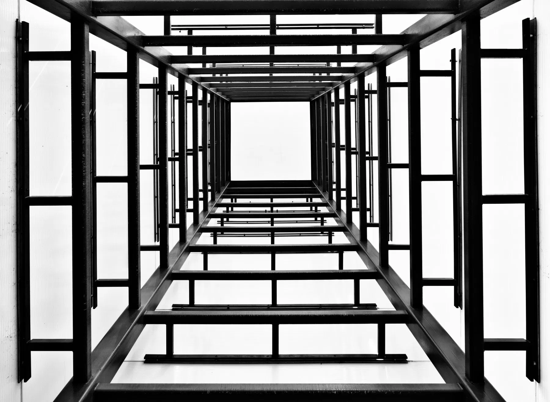 Black and white photograph looking straight up through a tall, open rectangular tower structure. Dark metal or steel beams form repeating square frames that recede upward toward a bright white sky, creating a dramatic tunnel-like perspective. The geometric pattern of horizontal and vertical bars grows progressively smaller toward the center, drawing the eye to a bright square of light at the top.