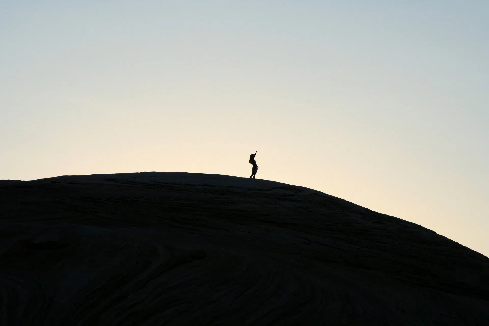 A lone figure silhouetted against a pale dusk sky stands atop a large, smooth rock formation, one arm raised. The rock dominates the lower half of the image in near-black shadow, its surface texture faintly visible, creating a striking contrast with the soft gradient of the sky above.