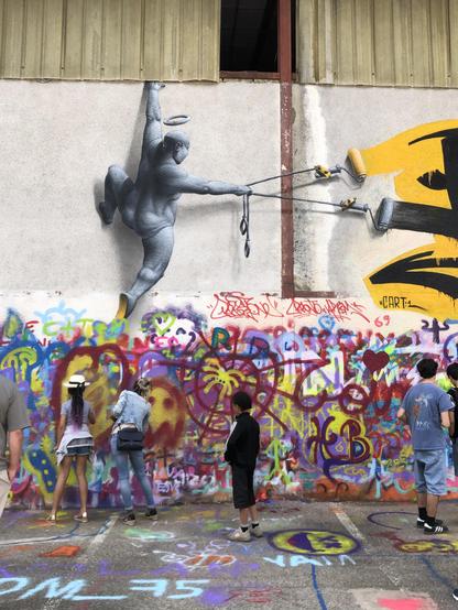 Au peinture fraiche festival (Bourgoin 38) le 9/05/26. Visiteurs s'initiant à l'art du tag sur les murs d'entrepôt désaffecté. Sur le haut dur mur un tag représentant un tagueur qui tire ses deux couleurs dans une position acrobatique...