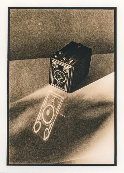 A vintage box camera rests on a sunlit surface, casting a striking elongated shadow. The strong sunlight also reflects off the camera’s glossy front panel onto the white glossy surface in front of it, creating a bright, ghostly reflection of the camera and its lens. The hard light emphasizes the camera’s geometric form and creates an interplay of solid black shapes, pale highlights, and reflected light. The lith printing process adds a warm sepia tone, coarse grain, and a slightly distressed, atmospheric texture, giving the image an aged, almost dreamlike quality.