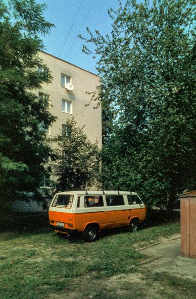 Red and white vintage Volkswagen Transporter van parked on a patch of grass in a residential courtyard. Tall green trees frame a pale five-story apartment block with a satellite dish on its wall, and a rusty brown shed stands at the right edge. Film photograph with visible grain and cool, blue-green tones.