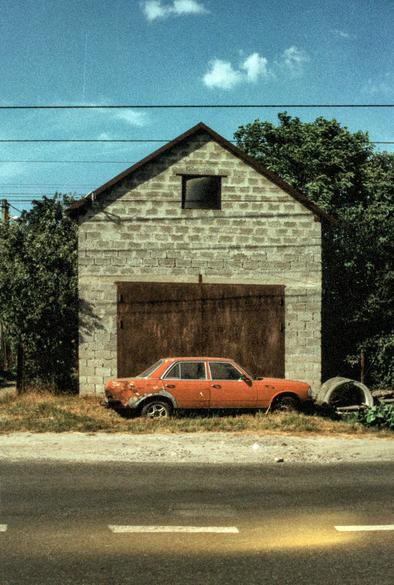 Rusty red vintage sedan with peeling paint parked on dry grass in front of a gray cinder-block garage with a large brown metal door. Power lines cut across a bright blue sky with a few small clouds, and green trees rise on both sides. An empty asphalt road with white dashed lines fills the foreground. Grainy film photograph with saturated colors.