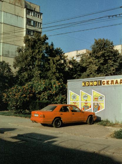 Red Mercedes sedan parked at an angle against a gray kiosk painted with a comic-style mural of three superheroes in yellow panels, under a sign reading "КОНФІСКЛАД". Green trees and Soviet-era apartment blocks rise behind, with dense overhead wires crossing a deep blue sky. Strong late-day light casts long shadows across the foreground. Film photograph with warm tones and visible grain.