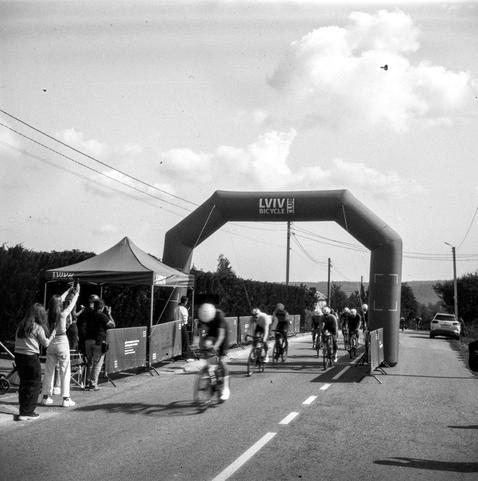 Black-and-white photo of road cyclists approaching a finish line under a large inflatable arch branded “Lviv Bicycle Club”. A blurred lead rider in a white helmet is closest to the camera, with a small group of riders close behind. On the left, spectators stand by a branded tent and barriers, and two women raise their phones to film the finish. Power lines cross a cloudy sky, and a parked car and hedges line the road.
