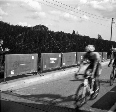 Black-and-white photo of a cyclist in motion blur riding past a line of banner-covered barriers along a hedge-lined road. The first cyclist crosses the finish line while the second, riding behind him, is partly visible at the right edge. A spectator on the left, wearing sunglasses, applauds. Overhead wires and a guy-wire cut diagonally across a cloudy sky.