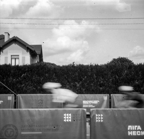Black-and-white photo of two blurred cyclists passing behind rows of barriers with Ukrainian-language sponsor banners. The riders appear as pale, streaked shapes in front of a tall dark hedge. A house with a gabled roof and chimney rises behind the hedge, and horizontal power lines run across a cloudy sky. The banner text in the foreground is sharp in contrast to the motion-blurred riders.