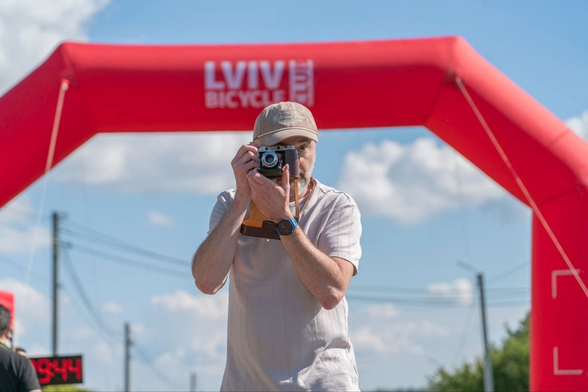 A man stands beneath a large red inflatable arch marked “Lviv Bicycle,” holding a vintage camera up to his face and taking a photograph. He wears a beige baseball cap, a light grey pinstriped T-shirt, and a wristwatch with a blue strap. A leather camera strap hangs around his neck. The bright blue sky, scattered white clouds, and utility wires in the background suggest a sunny day outdoors. The low angle and bold red arch frame the photographer, capturing a moment of concentration during a cycling event.