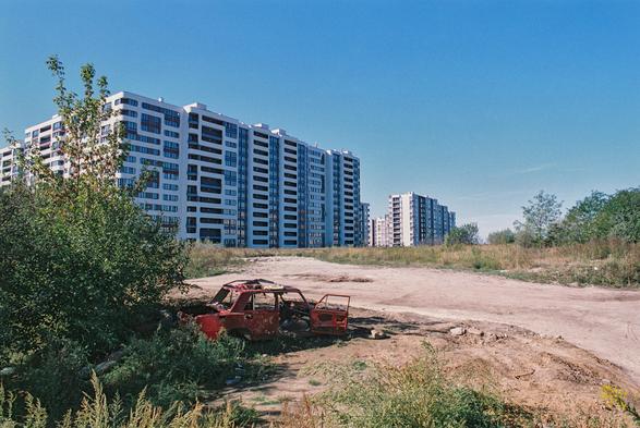 A sun-bleached dirt lot stretches beneath a clear blue sky, with a red car abandoned among weeds and overgrown bushes in the foreground. Its doors hang open, and its stripped interior contrasts with the bright, freshly built apartment blocks rising behind it. The harsh midday light, dry earth, and scattered rubble create a stark contrast between urban development and neglect