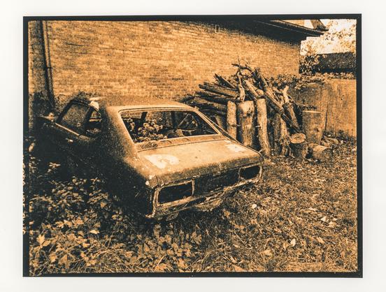 Lith print in warm amber and burnt-orange tones with deep black shadows and coarse grain, framed by a thick black border. An abandoned, rusting car sits at an angle in an overgrown yard, seen from the rear. Its paint is peeling, the tail lights are missing, and the rear window is open to the weeds growing inside. Fallen leaves and wild plants surround it. Behind it are a brick wall and a stack of cut logs.